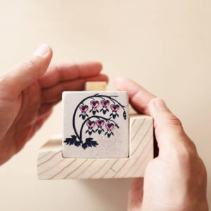 Nahaufnahme des Namenssteins von ÜHolz® Jonny mit eingraviertem Motiv Tränendes Herz, verziert mit pinkfarbenen Glaskristallen. Zwei Hände halten den Stein sanft – ein Symbol dafür, dass der Naturstein aus dem ÜHolz® herausnehmbar ist und auch zu Hause als Erinnerungsstück einen würdevollen Platz findet.